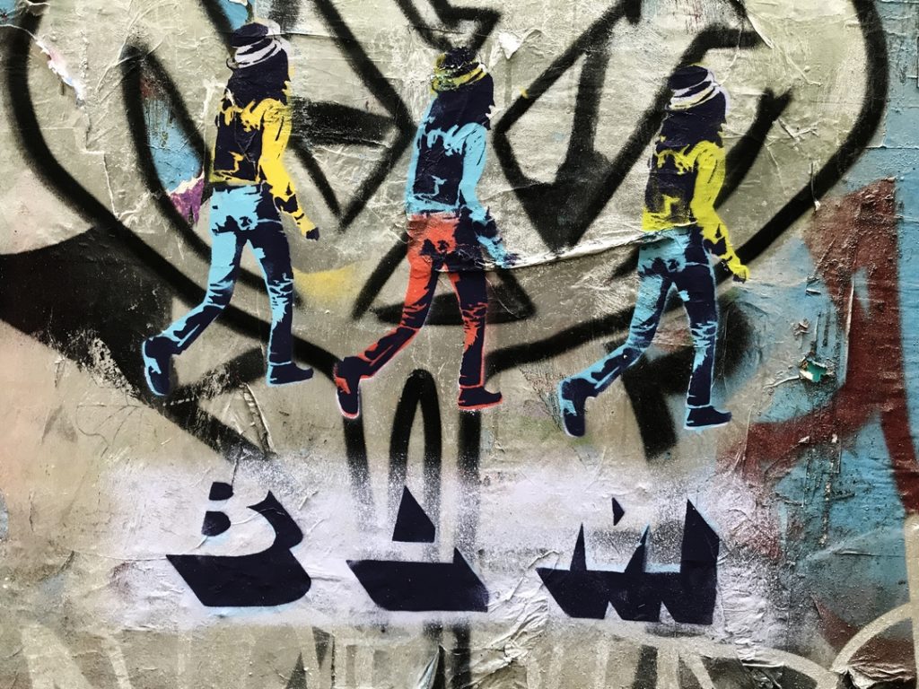 Stencil street art of 3 figures waling into the background wearing bright coloured clothing with letters BLM below them