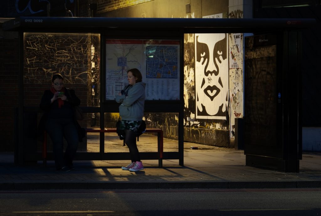 Huge Obey Giant paste up in Shoreditch seen through a bus shelter at night with people patiently waiting for bus