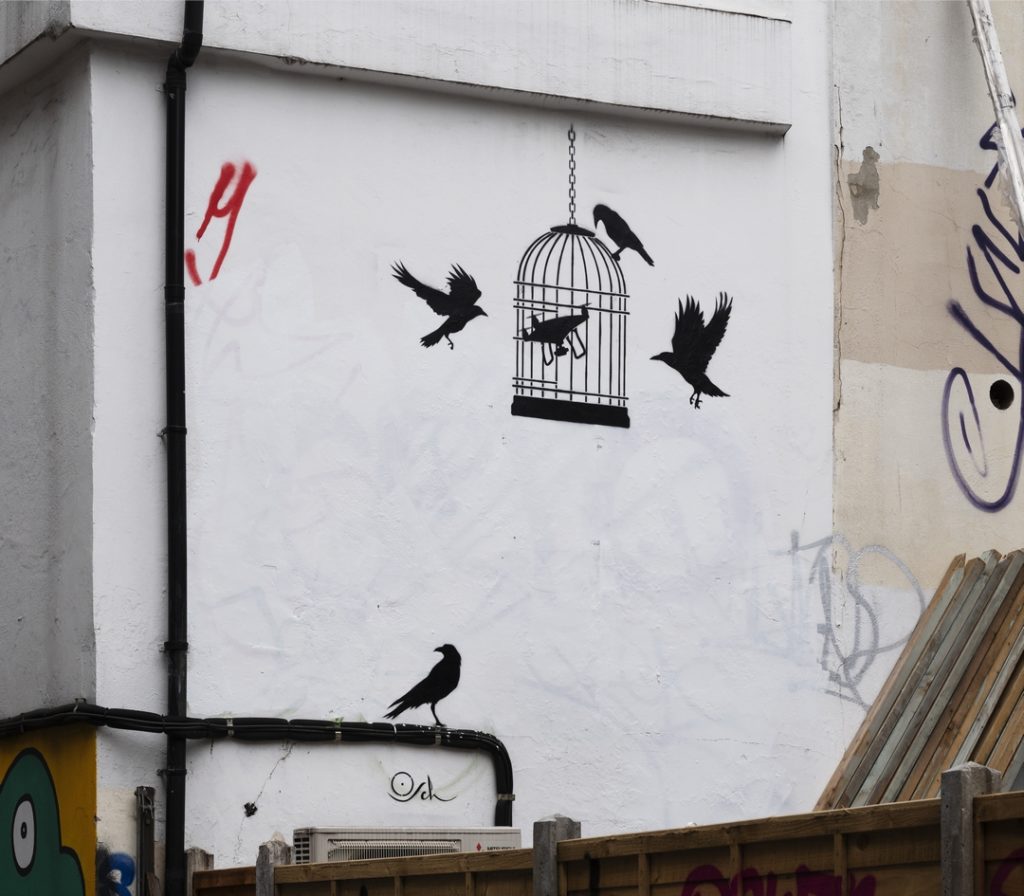 Street art stencil onside of a building shoing a drone inside a birdcage with crows pecking outside the cage, by street artist Otto Schade, called "Artificial Intelligence"